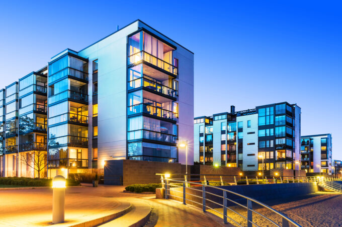 Modern apartment buildings with large glass windows, balconies, and outdoor lights illuminate the area at dusk. A curved walkway with railings leads past landscaped areas in the foreground.