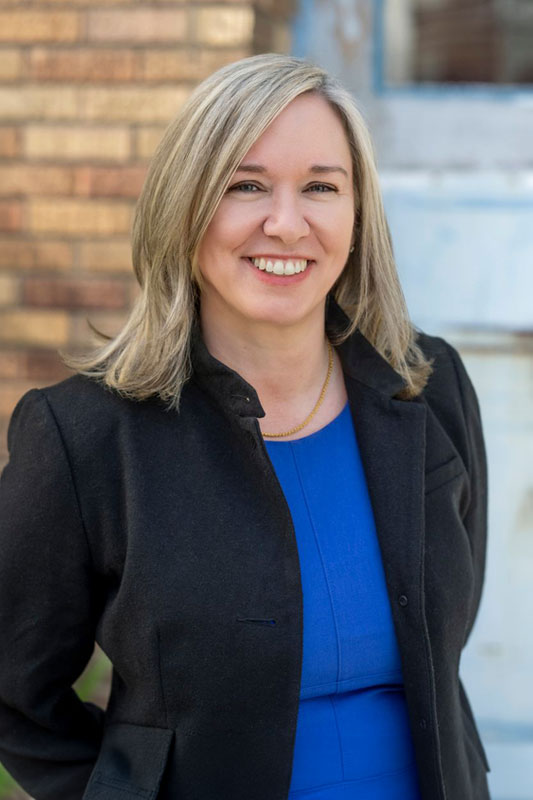 A Woman With Blonde Hair Wearing A Black Blazer Over A Blue Dress Is Smiling While Standing Outdoors In Front Of A Brick Wall.