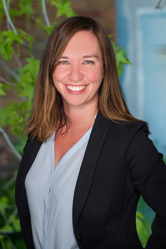 A Smiling Woman With Straight Brown Hair Wearing A Black Blazer Over A Light Blue Blouse Stands In Front Of A Blurred Background With Green Leaves And A Brick Wall.