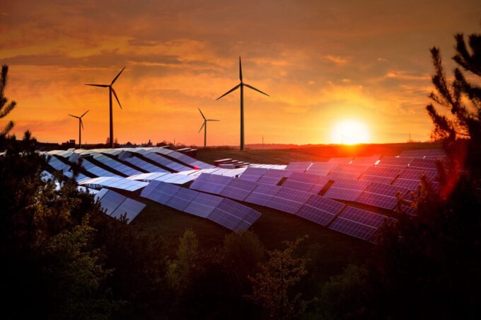 Rows of solar panels and wind turbines stand in a field at sunset, with the sky glowing orange and the sun low on the horizon, surrounded by trees and greenery.